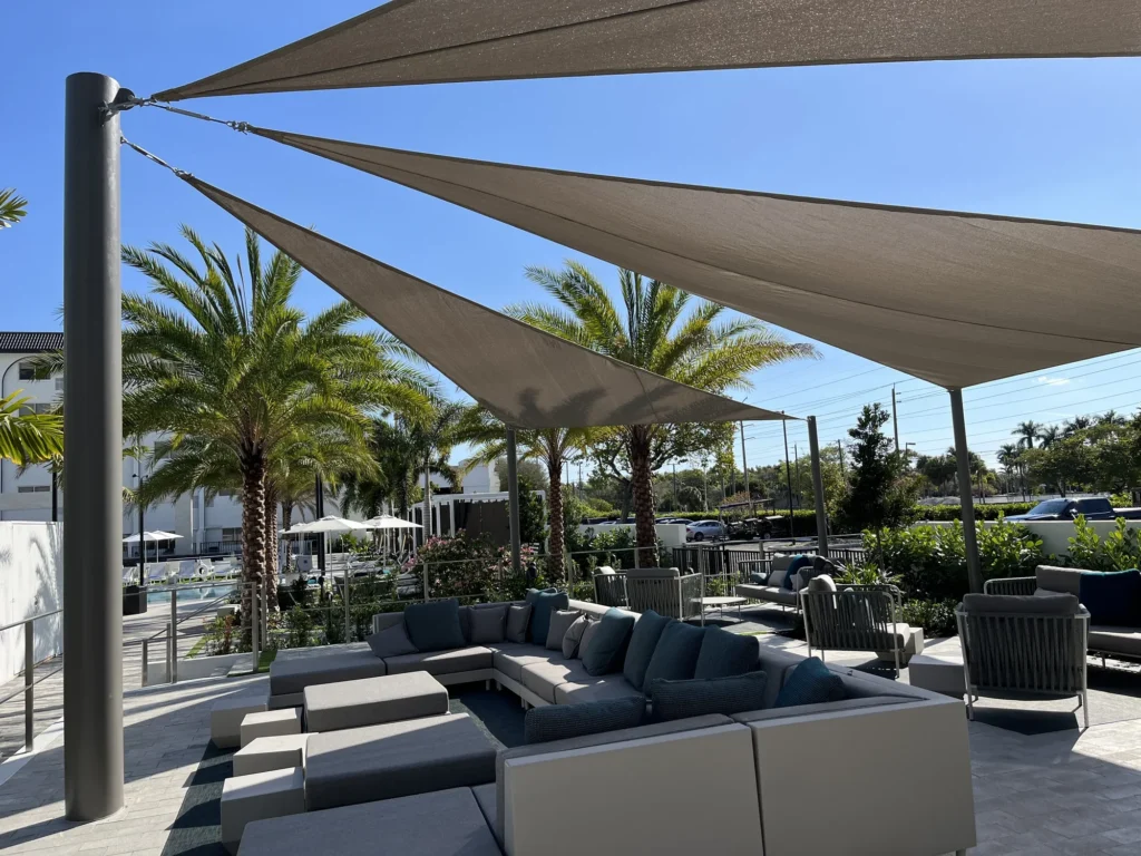 Overlapping shade sails above terrace sofas beside a white building and palm trees.